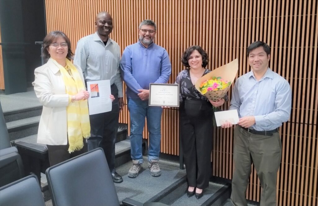 5 people are shown standing side by side in a lecture hall at the University of Lethbridge. From the left, Glenda Bonifacio is shown smiling and wearing a yellow scarf and holding information about Habiba Kadiri's book launch. Next from the left, Olu Awosoga is shown smiling and standing next to Saurya Das, who is also smiling. Next from teh left, Habiba Kadiri is shown smiling and holding a certificate and a bouquet of flowers. And finally,to the left of Habiba is Gideon Fujiwara, shown smiling, who is also holding a piece of paper.